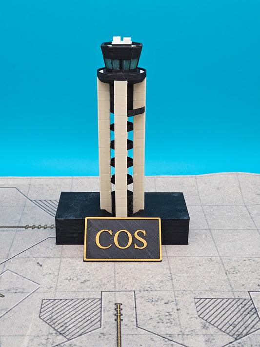 A miniature model of an air traffic control tower with a black base and a sign reading 'COS' in gold lettering, set against a bright blue background and a gray tiled surface.