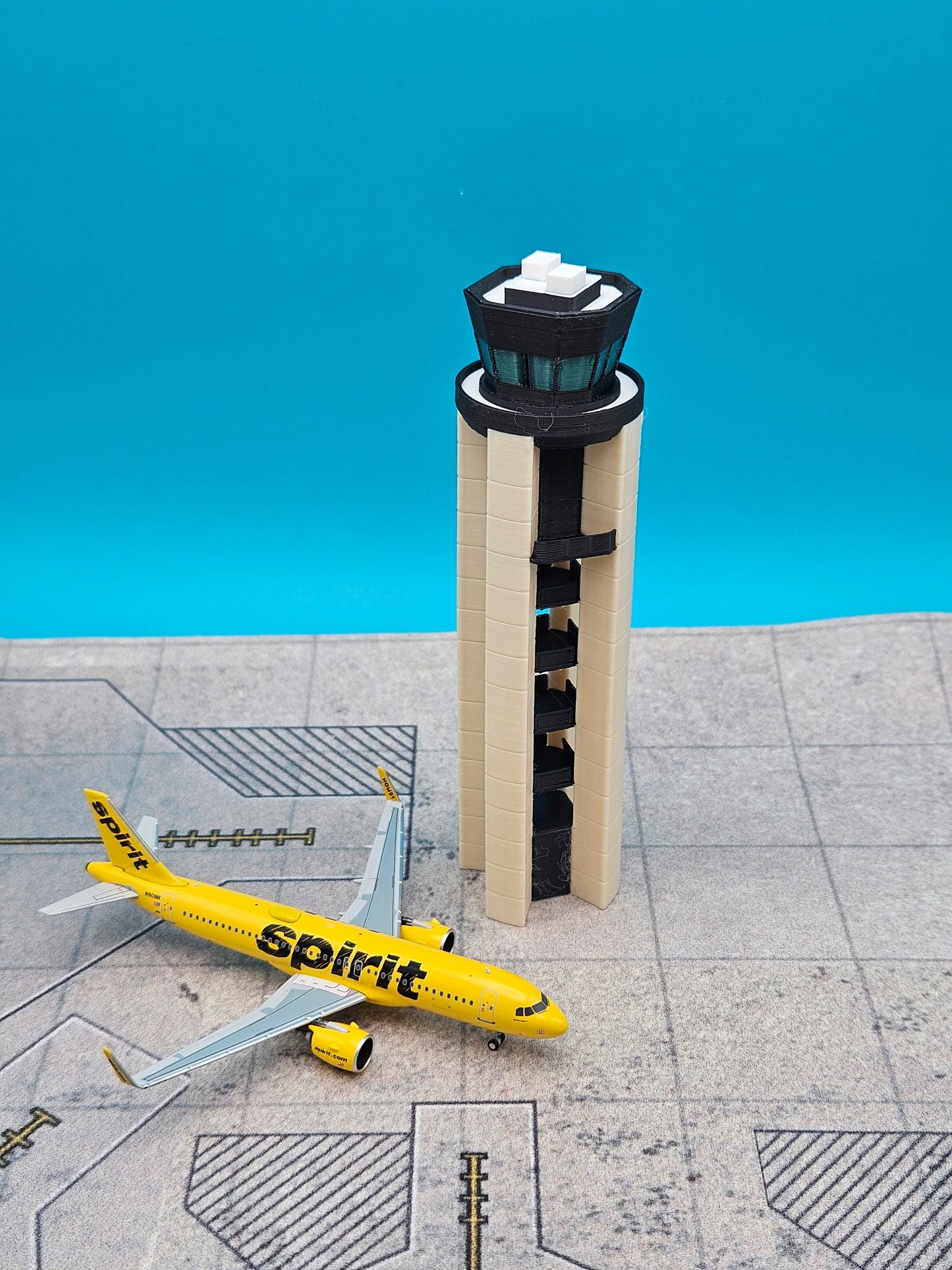 A miniature model of a yellow Spirit Airlines airplane next to a beige and black airport control tower, displayed on a gray runway pattern against a bright blue background.
