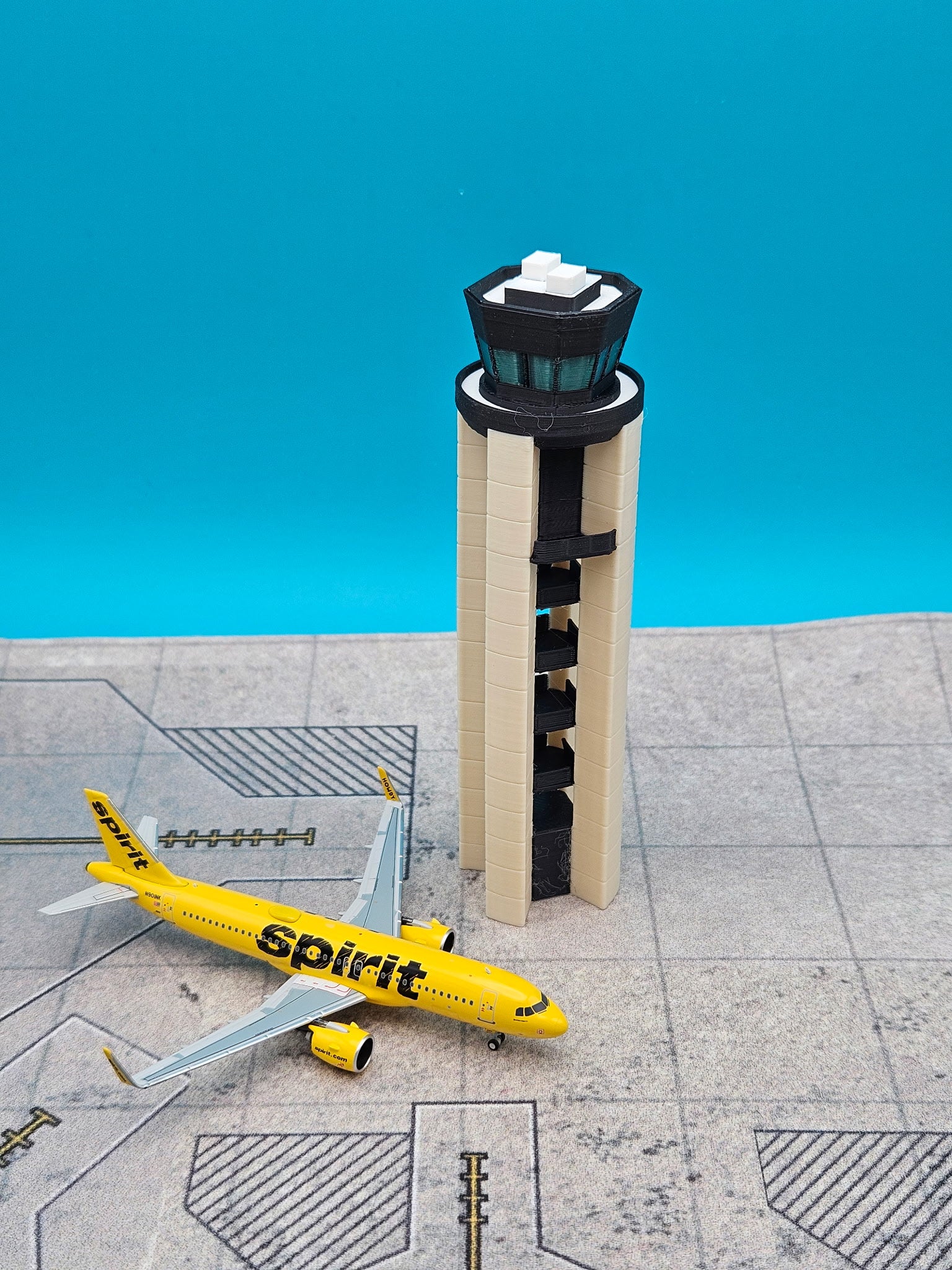 A miniature model of a yellow Spirit Airlines airplane next to a beige and black airport control tower, displayed on a gray runway pattern against a bright blue background.