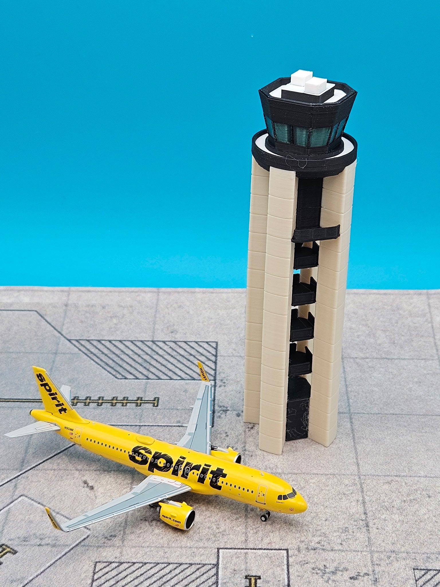 A miniature model of a yellow Spirit Airlines airplane next to a beige and black airport control tower, displayed on a gray runway surface against a bright blue background.
