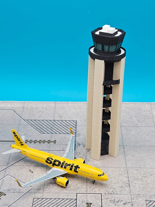 A miniature model of a yellow Spirit Airlines airplane next to a beige and black airport control tower, displayed on a gray runway surface against a bright blue background.