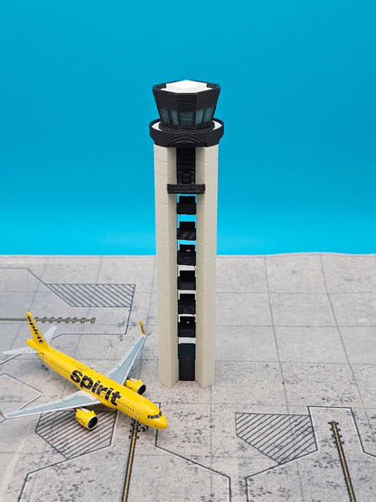 A miniature model of an airport control tower stands next to a yellow Spirit airplane on a gray tiled runway surface against a bright blue background.