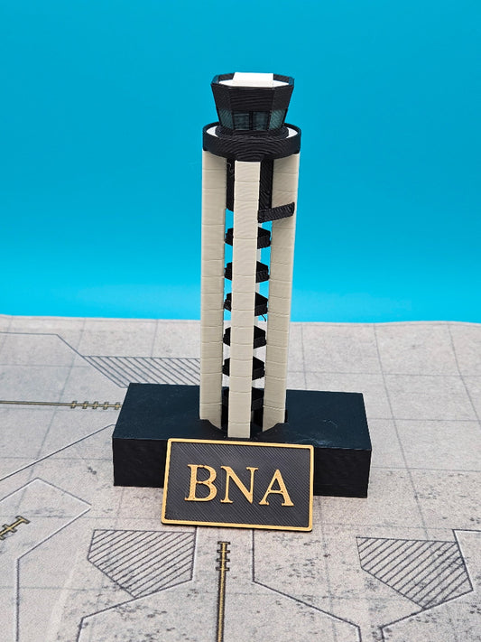 A miniature model of the BNA (Bogota National Airport) tower, built from interlocking toy blocks, standing on a black base with a gold sign that reads 'BNA'. The model is set against a bright blue backdrop on a gray patterned surface.
